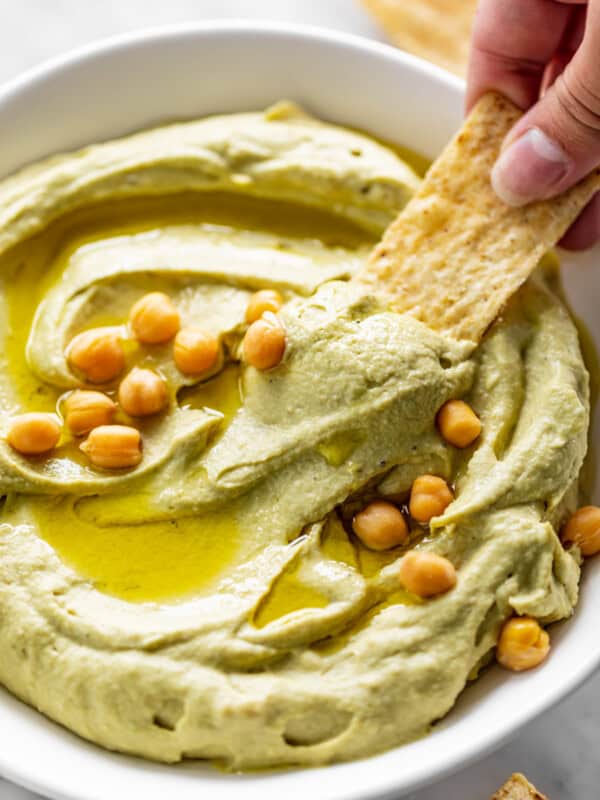 A corn chip dips into a white bowl full of Avocado Hummus, drizzled with olive oil and topped with whole chickpeas.
